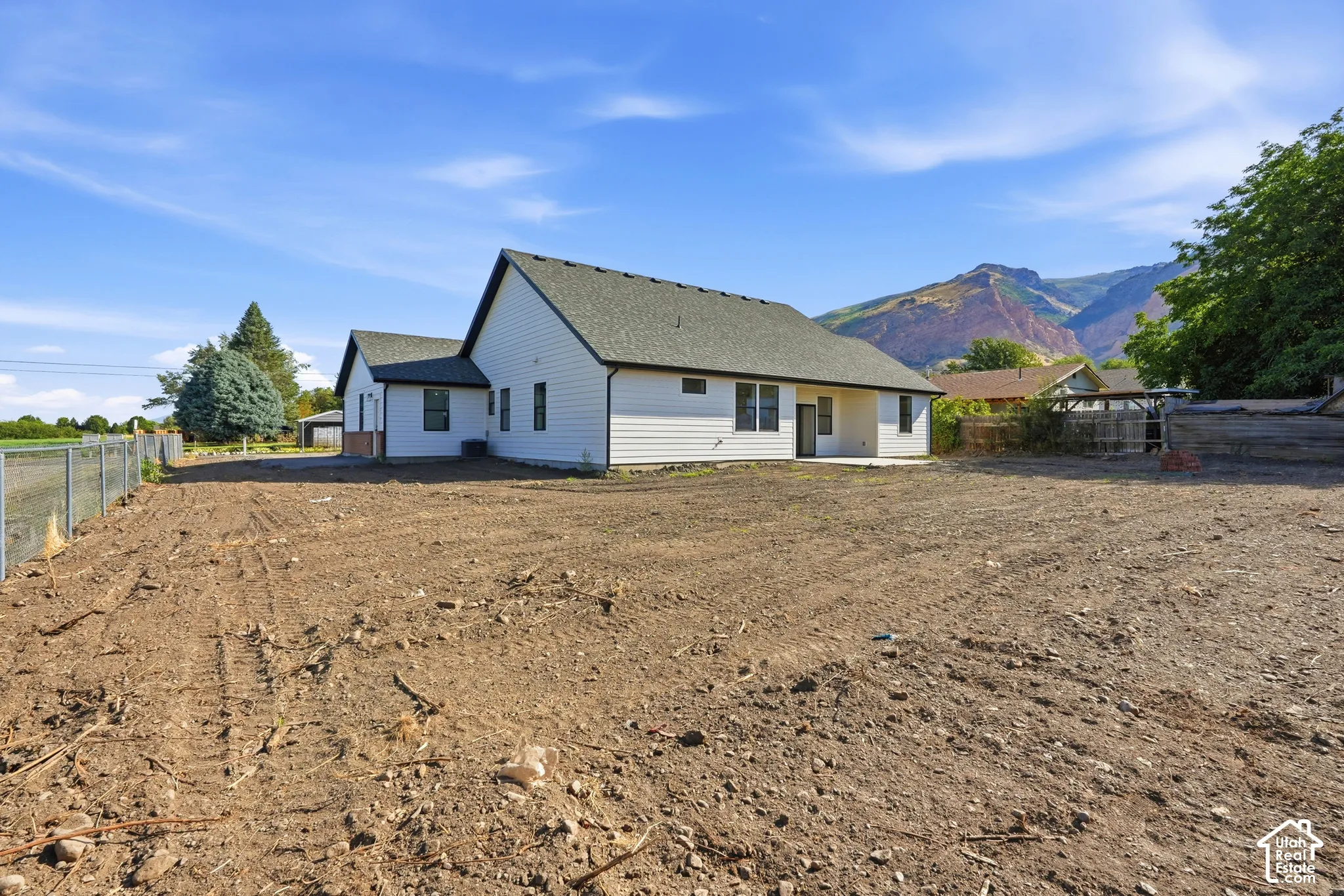 Expansive undeveloped lot with chain-link fencing, featuring a single-story residence with white siding, dark trim, and a patio area