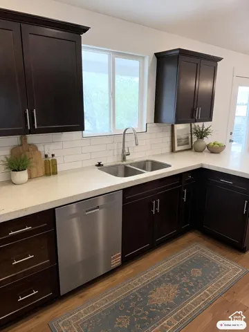 Kitchen featuring dark wood-finish cabinetry, light-toned countertops, a subway tile backsplash, a stainless steel double basin sink with a gooseneck faucet, and a stainless steel dishwasher