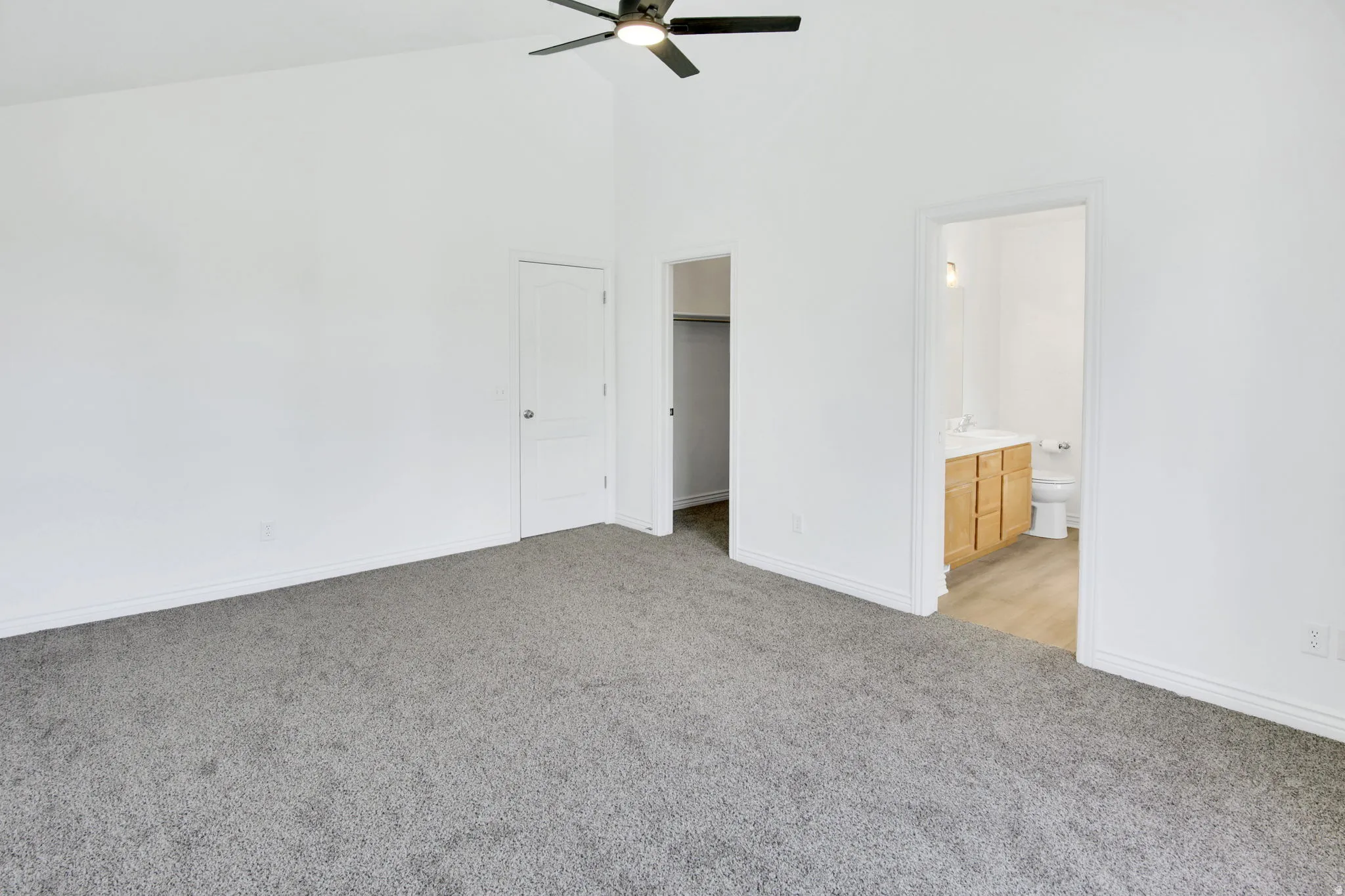 Spacious room featuring vaulted ceilings, a ceiling fan, grey carpeting, a white paneled door, and an entryway to a bathroom with a wood-finish vanity and hard-surface flooring