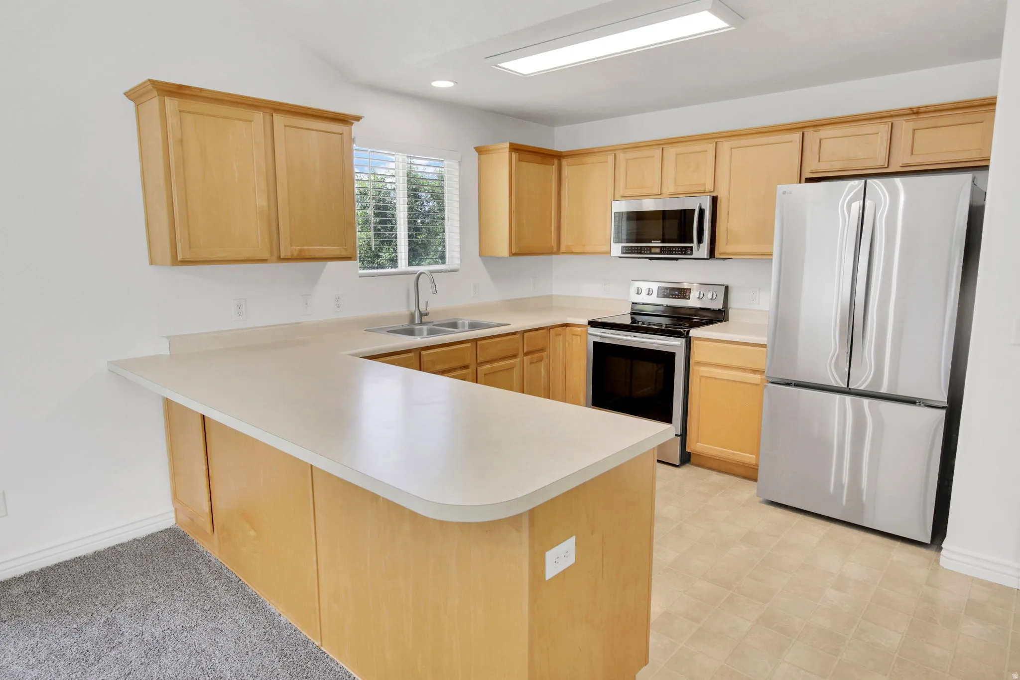 Kitchen featuring light wood-finish cabinetry, stainless steel appliances, a double basin sink, light-toned countertops, and vinyl flooring