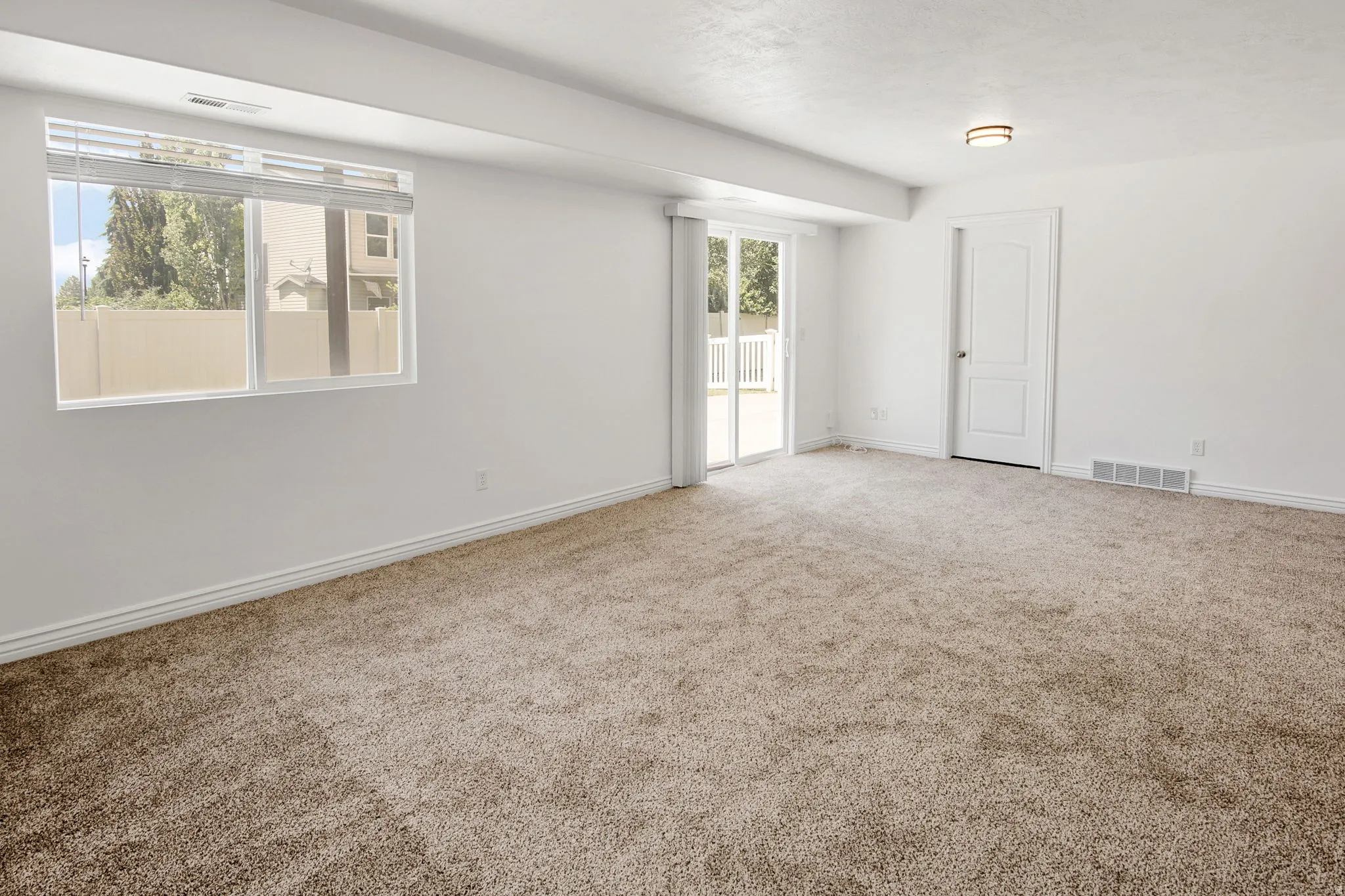 Spacious room featuring neutral carpeting, white painted walls, a large window with blinds, a sliding glass door to an exterior space, and a white interior door