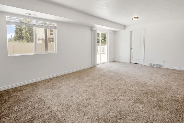 Spacious room featuring neutral carpeting, white painted walls, a large window with blinds, a sliding glass door to an exterior space, and a white interior door