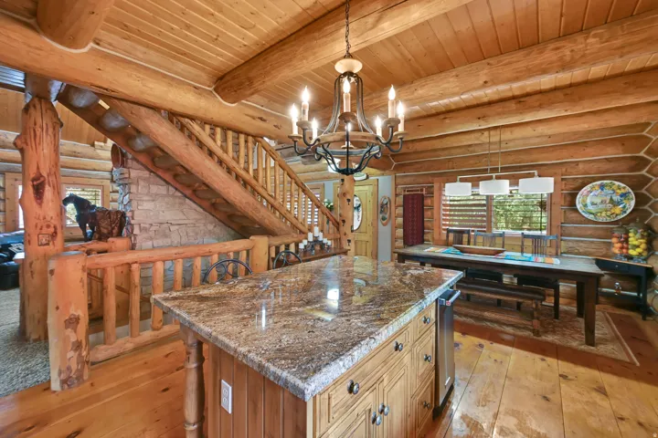 Open-concept interior featuring prominent exposed log walls and ceiling beams, a central kitchen island with a granite countertop, wood-finish flooring, and a log staircase with a stone-clad base