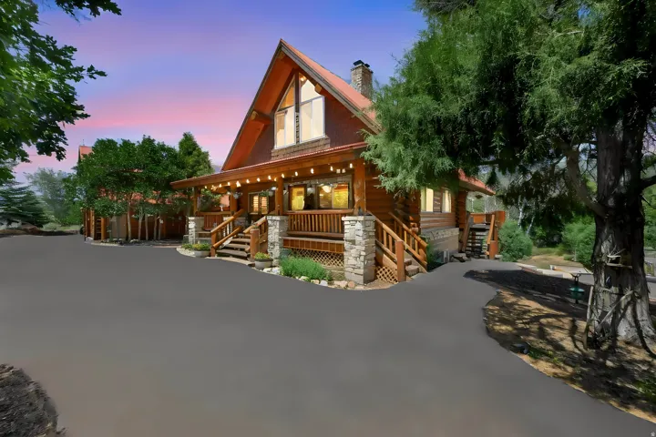 Rustic log home exterior featuring a stone chimney, covered porch with string lighting, and a paved driveway