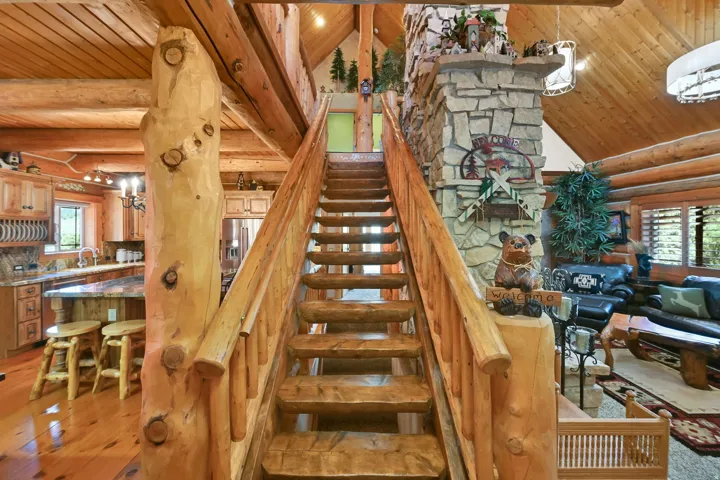 Rustic log staircase with natural wood finishes, a prominent stone fireplace, wood-finish flooring, and a kitchen featuring light wood cabinetry