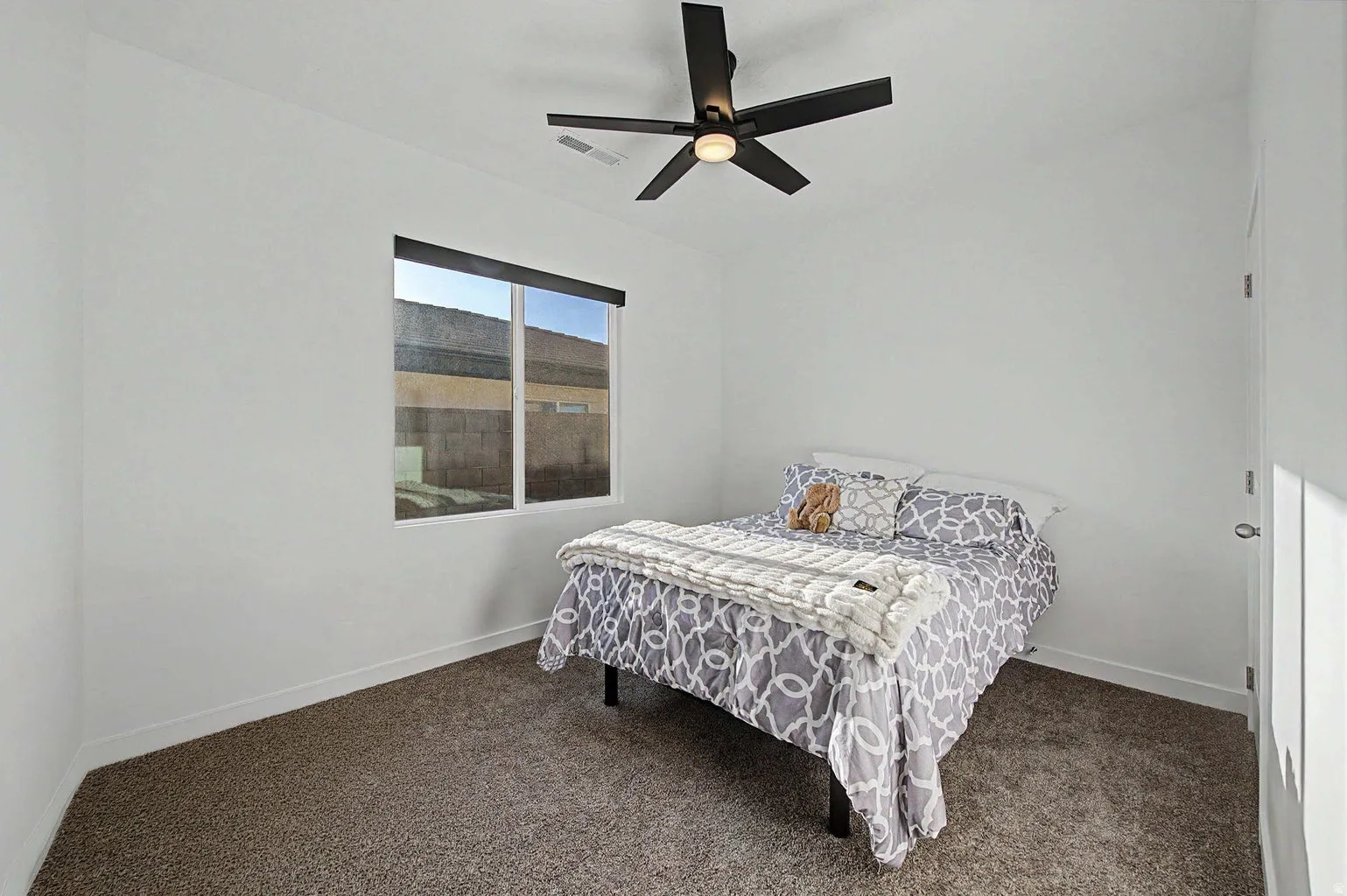 White walls with a ceiling-mounted fan, a window with a black roller shade, and plush carpet flooring