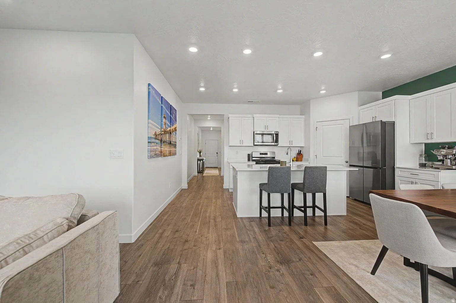 Open-concept living area featuring wood-finish flooring, a kitchen with white cabinetry, a central island with countertop seating, and stainless steel appliances