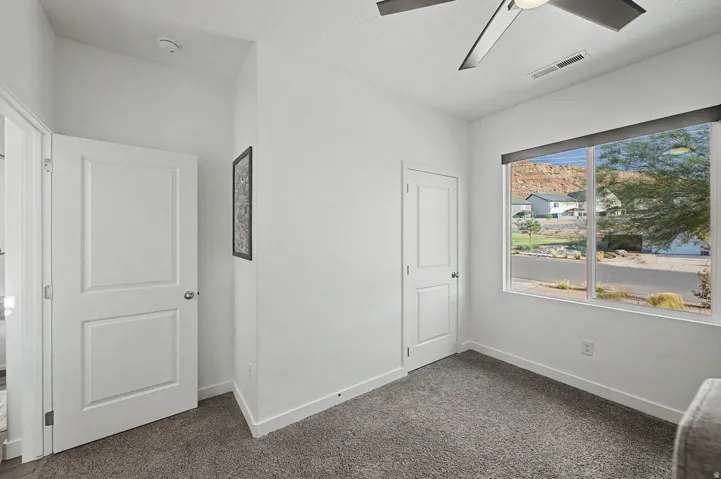 Bright room featuring a large window with roller shades, a ceiling fan with light fixture, and plush gray carpeting