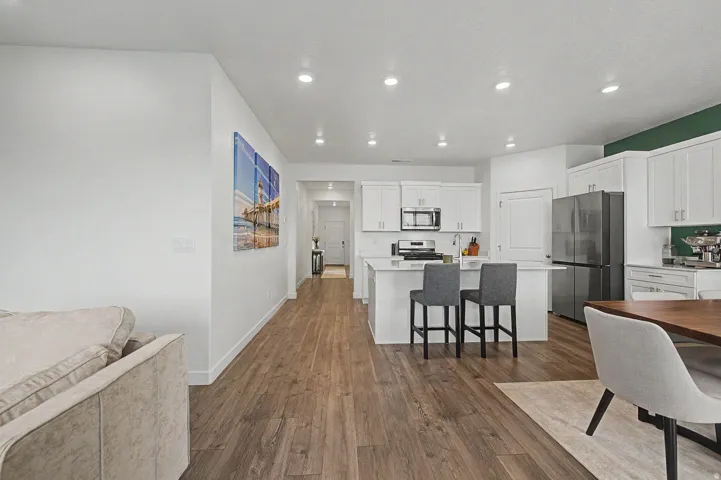Open-concept living area featuring wood-finish flooring, a kitchen with white cabinetry, a central island with countertop seating, and stainless steel appliances