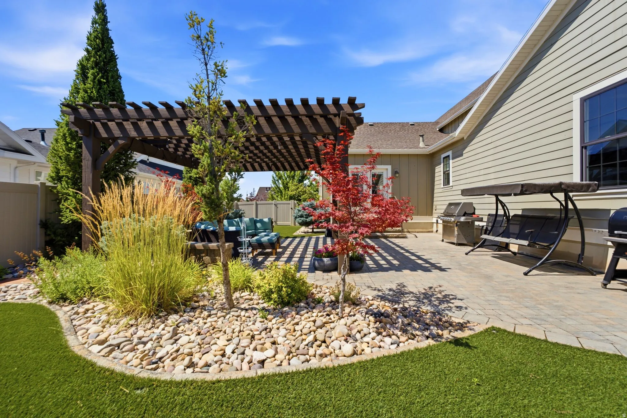 Expansive paver patio featuring a dark wood pergola, artificial turf, and decorative rock landscaping