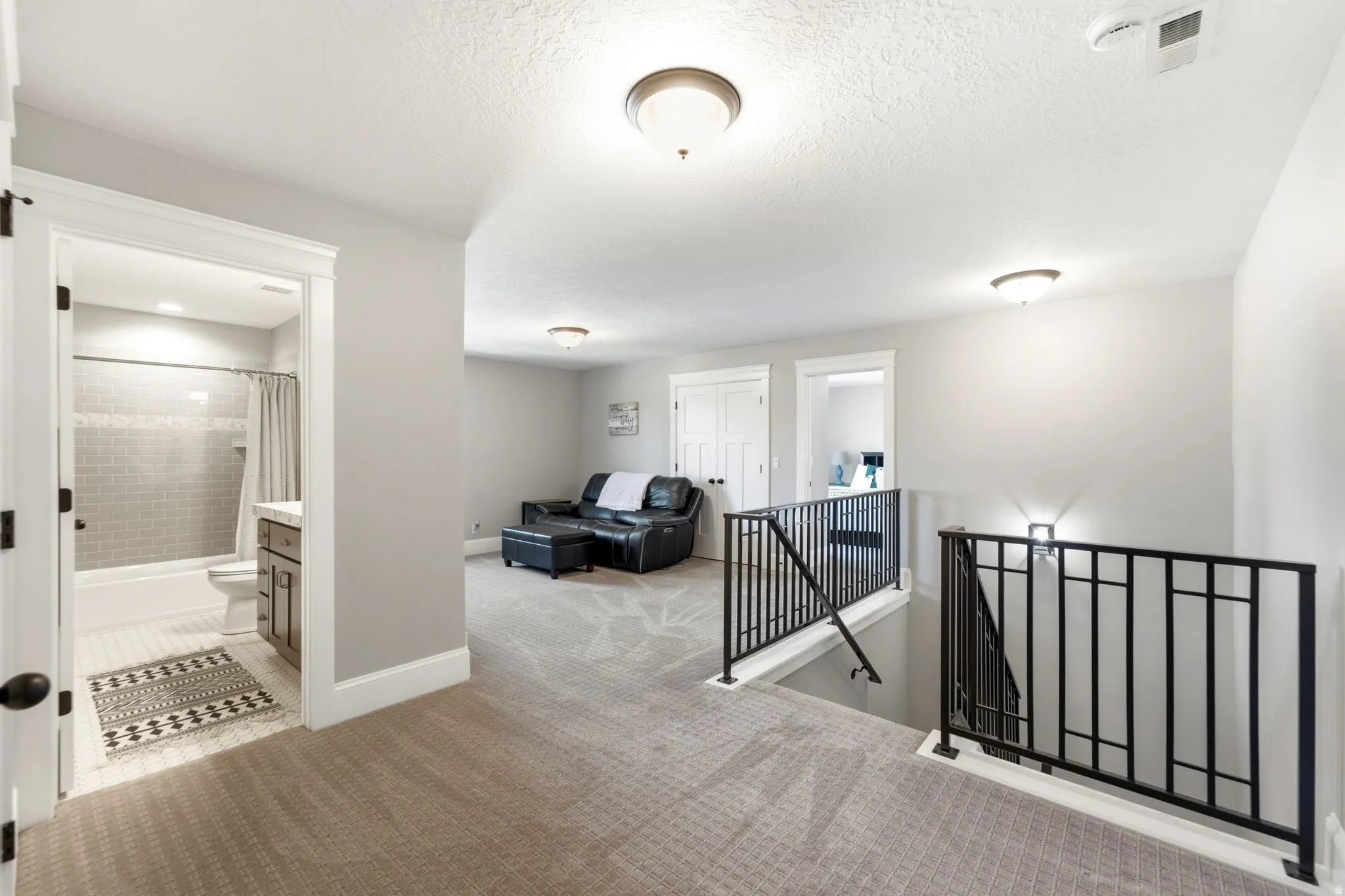 Upper-level landing featuring textured carpeting, a black wrought iron railing, and a full bathroom with a tiled shower and vanity