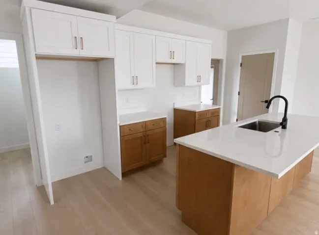 Contemporary kitchen featuring an expansive island with a stainless steel sink and matte black faucet, light wood-finish flooring, white upper cabinetry with gold-toned hardware, and a built-in refrigerator enclosure