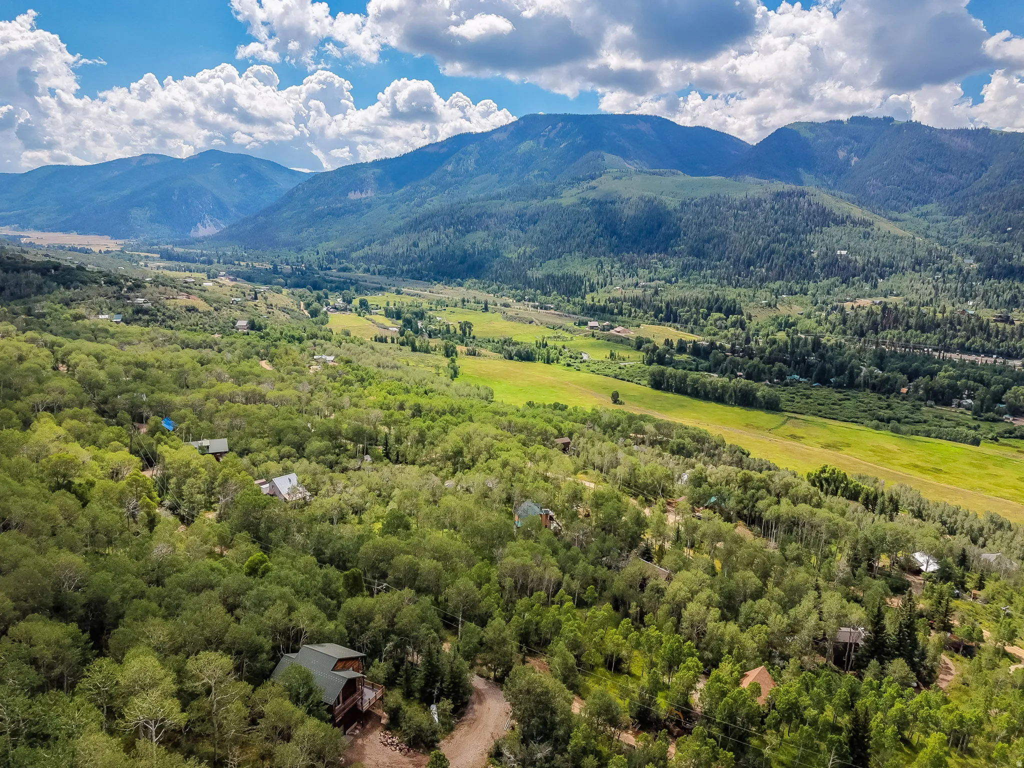 Expansive valley with verdant fields and dense tree coverage, bordered by distant mountains under a blue sky with white clouds