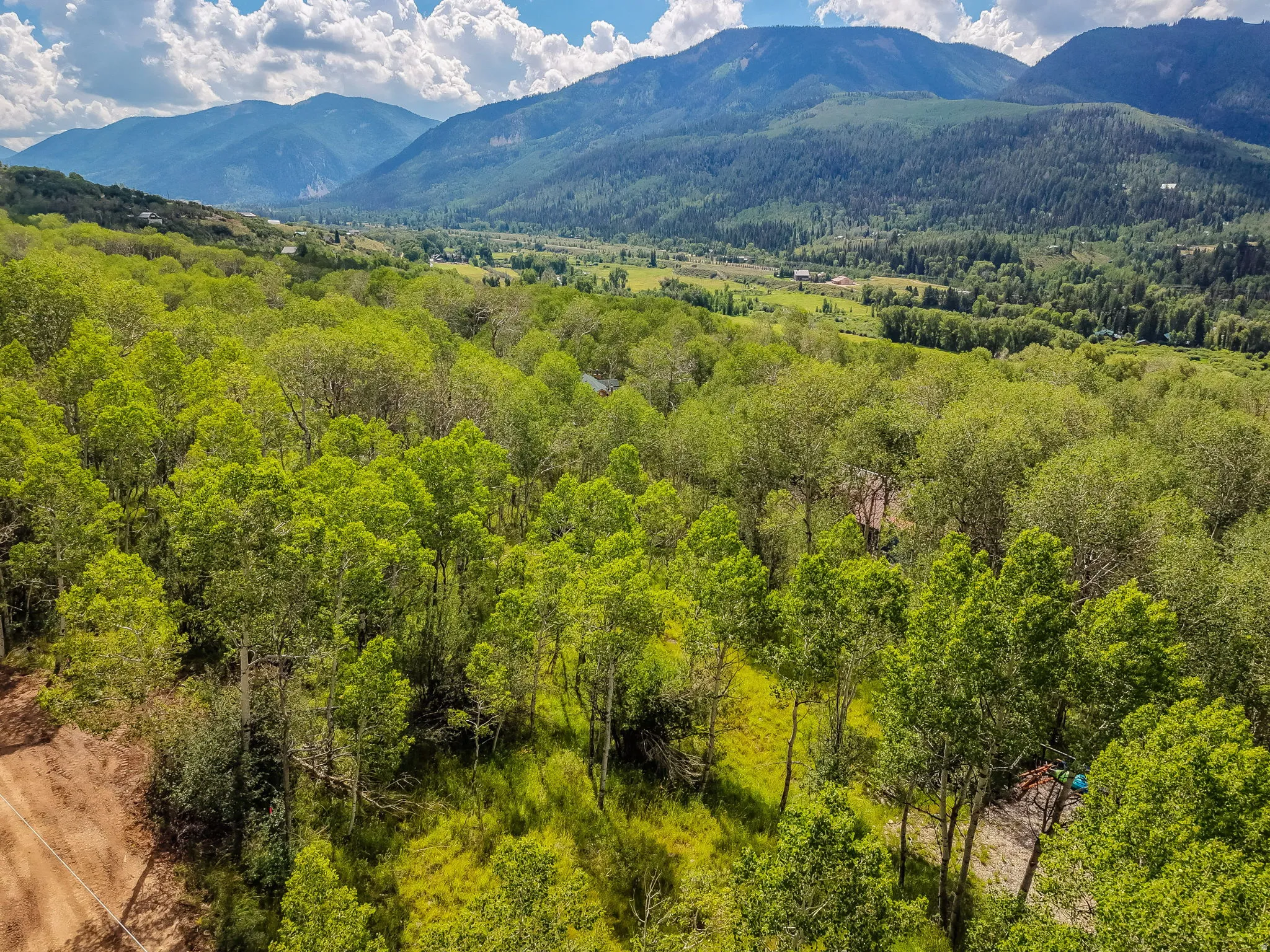 Expansive mountain landscape featuring a lush valley, dense tree coverage, and distant mountain ranges under a partly cloudy sky