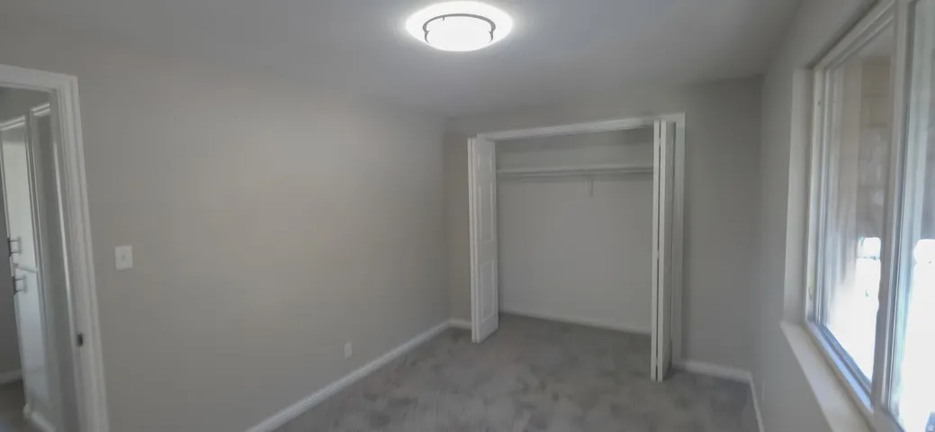 Room featuring light gray walls, gray carpet, a ceiling-mounted light fixture, a window with white trim, and a bi-fold closet with shelving