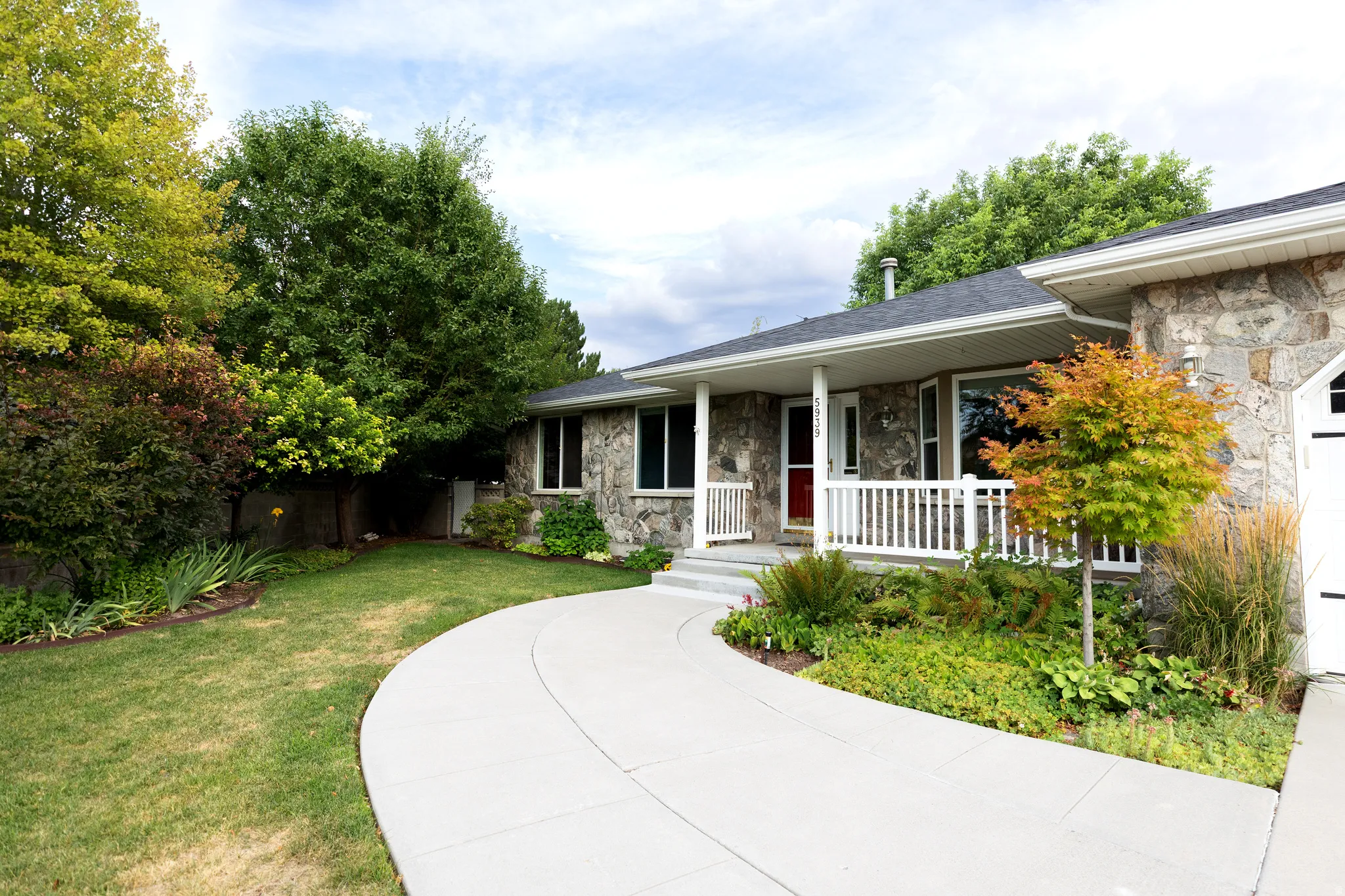 Stone-clad exterior featuring a covered porch with white railings, a concrete pathway, a red entry door, and mature landscaping