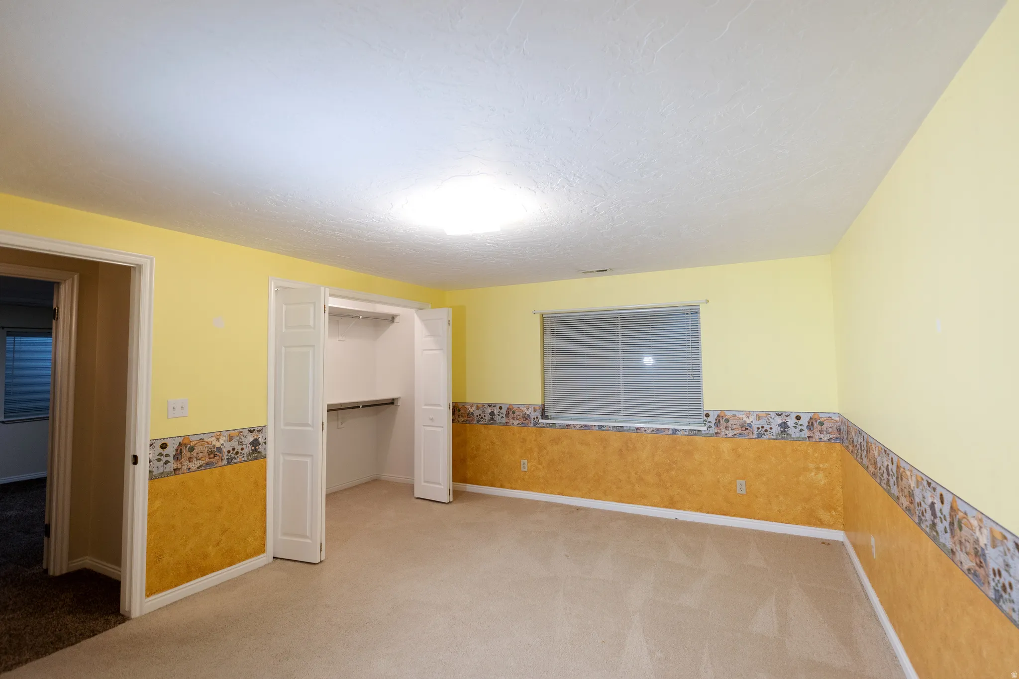 Spacious room featuring neutral carpeting, a large window with blinds, and an open closet with shelving and hanging rods