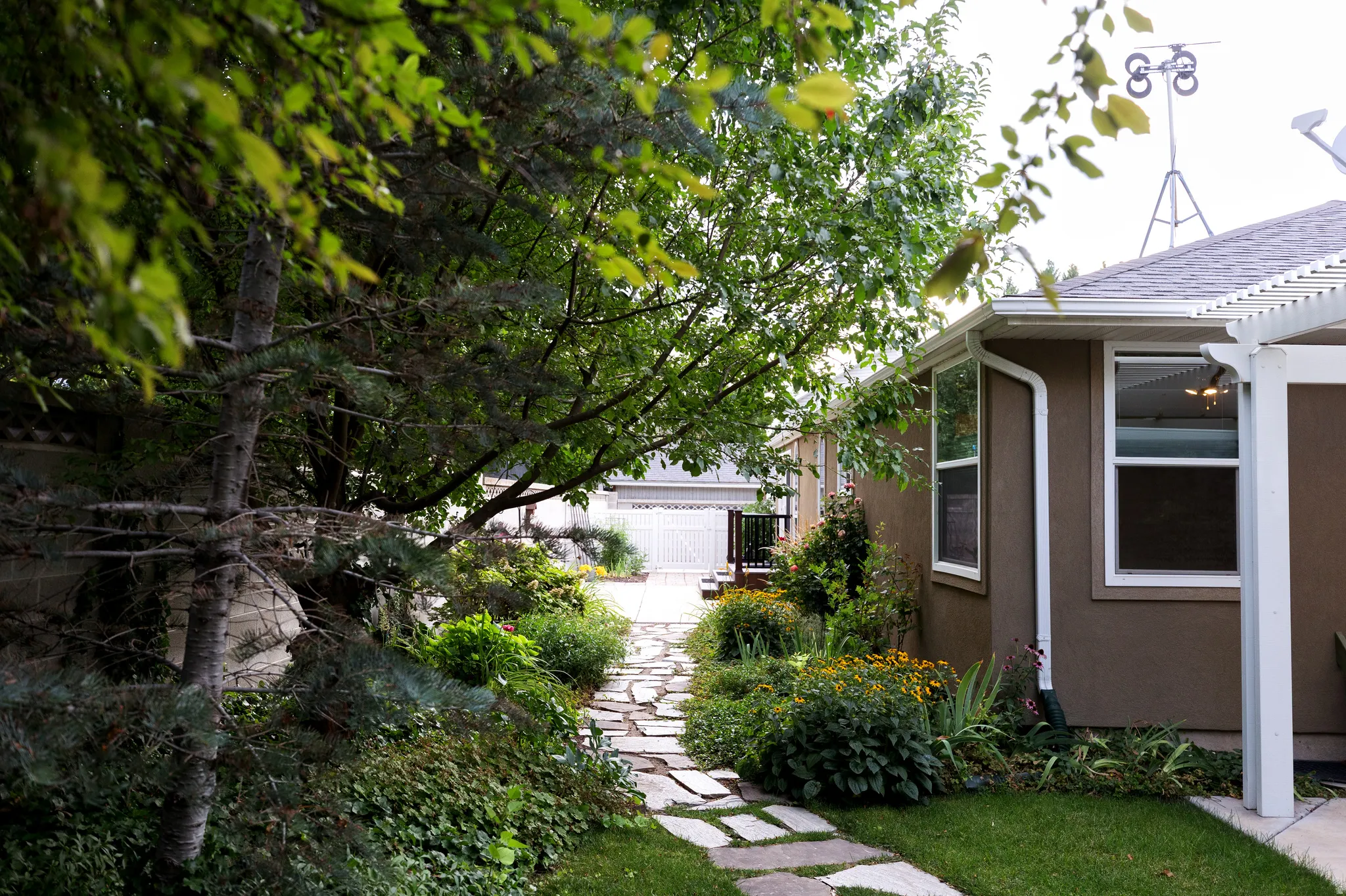 Landscaped side yard featuring a flagstone path, mature trees, and garden beds, bordered by a stucco exterior with white-framed windows and a white pergola