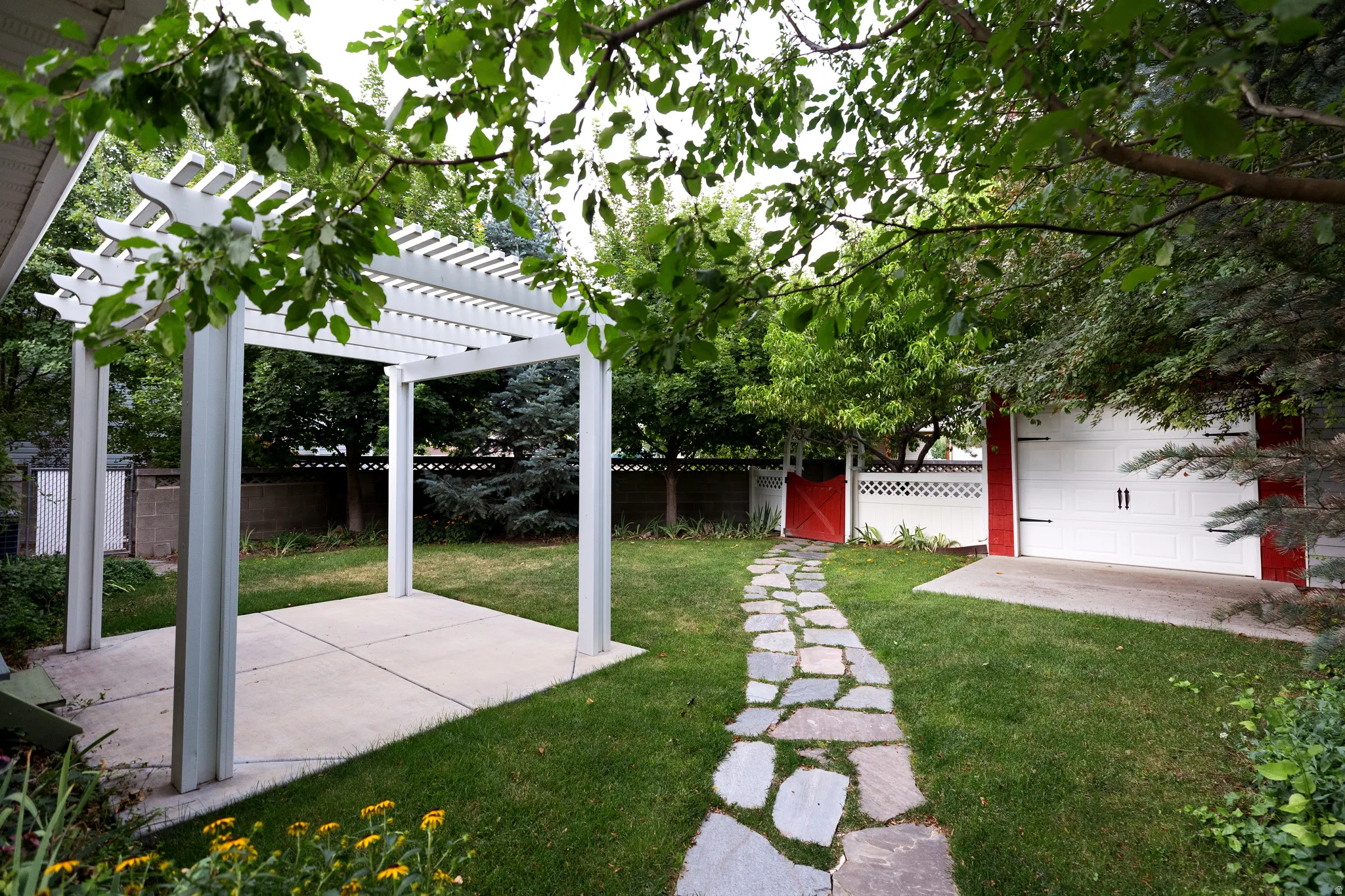 Expansive green lawn featuring a flagstone pathway, a white pergola over a concrete patio, a red painted garage with white doors, and a red gate