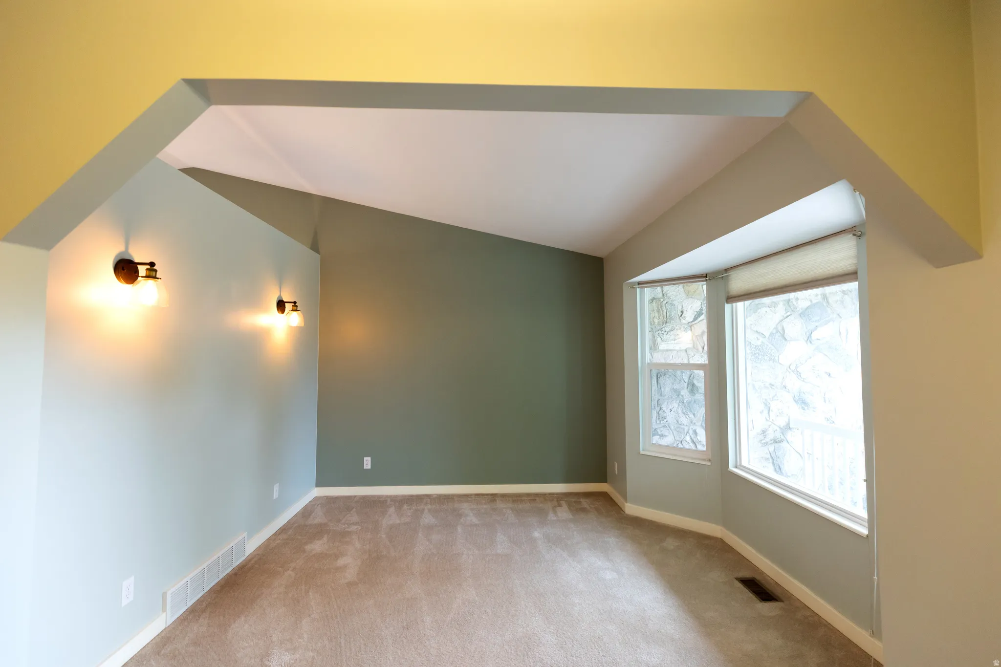 Carpeted room featuring a vaulted ceiling and a partial bay window with roller shades