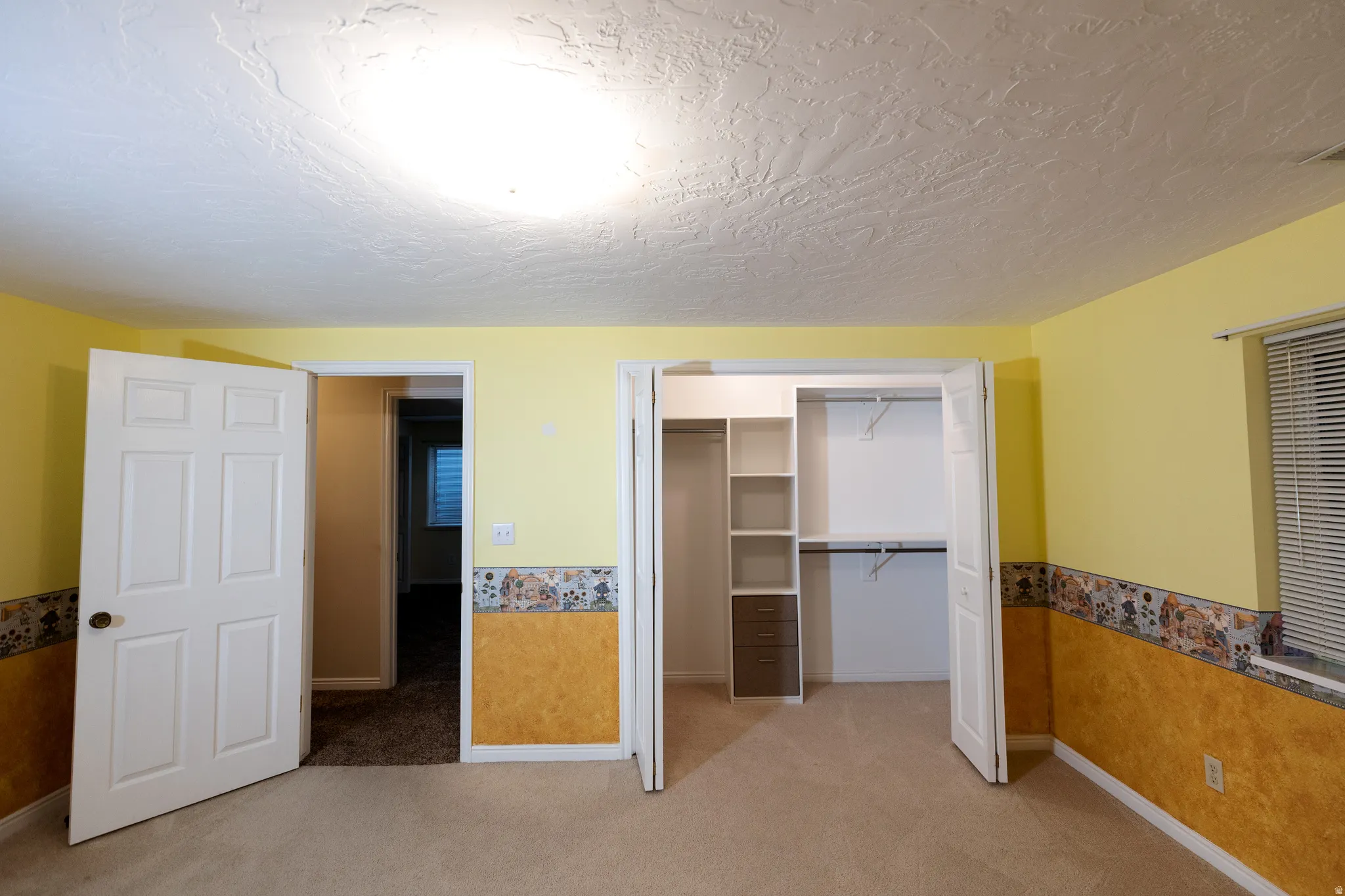 Room featuring light yellow walls with a decorative border and textured lower half, light beige carpet, white six-panel doors, and a built-in closet system with shelves and drawers