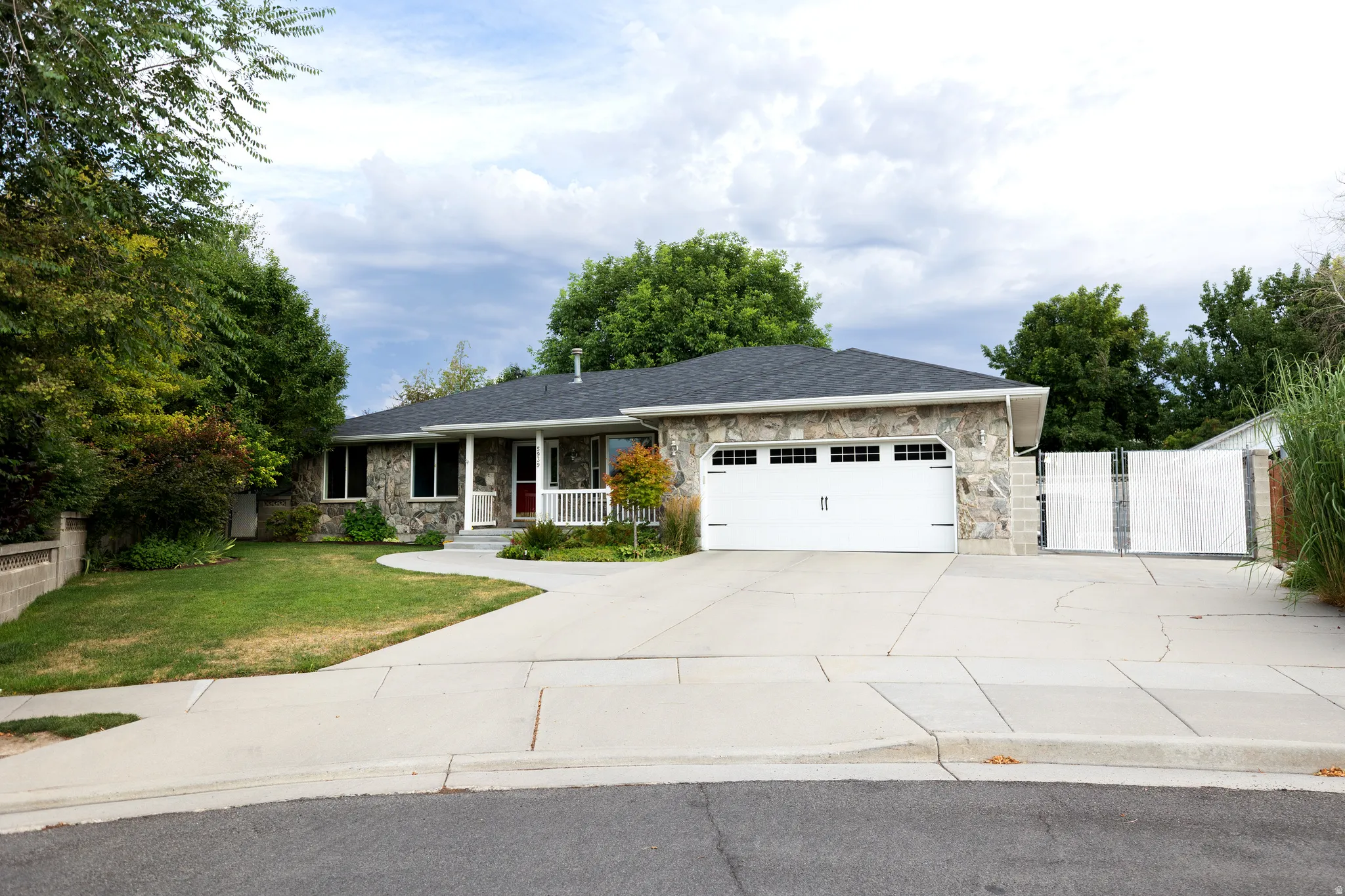Stone-clad ranch-style exterior featuring a large two-car garage with a paneled door, a covered front porch with white railings, and a concrete driveway