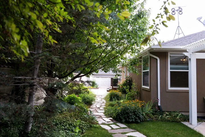 Landscaped side yard featuring a flagstone path, mature trees, and garden beds, bordered by a stucco exterior with white-framed windows and a white pergola