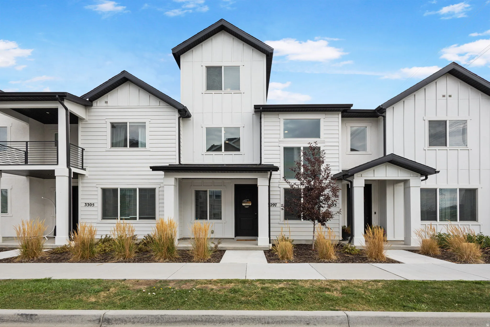 Modern farmhouse-style townhome featuring white siding with black trim, multiple gables, a covered entry, and a second-story balcony with black railings