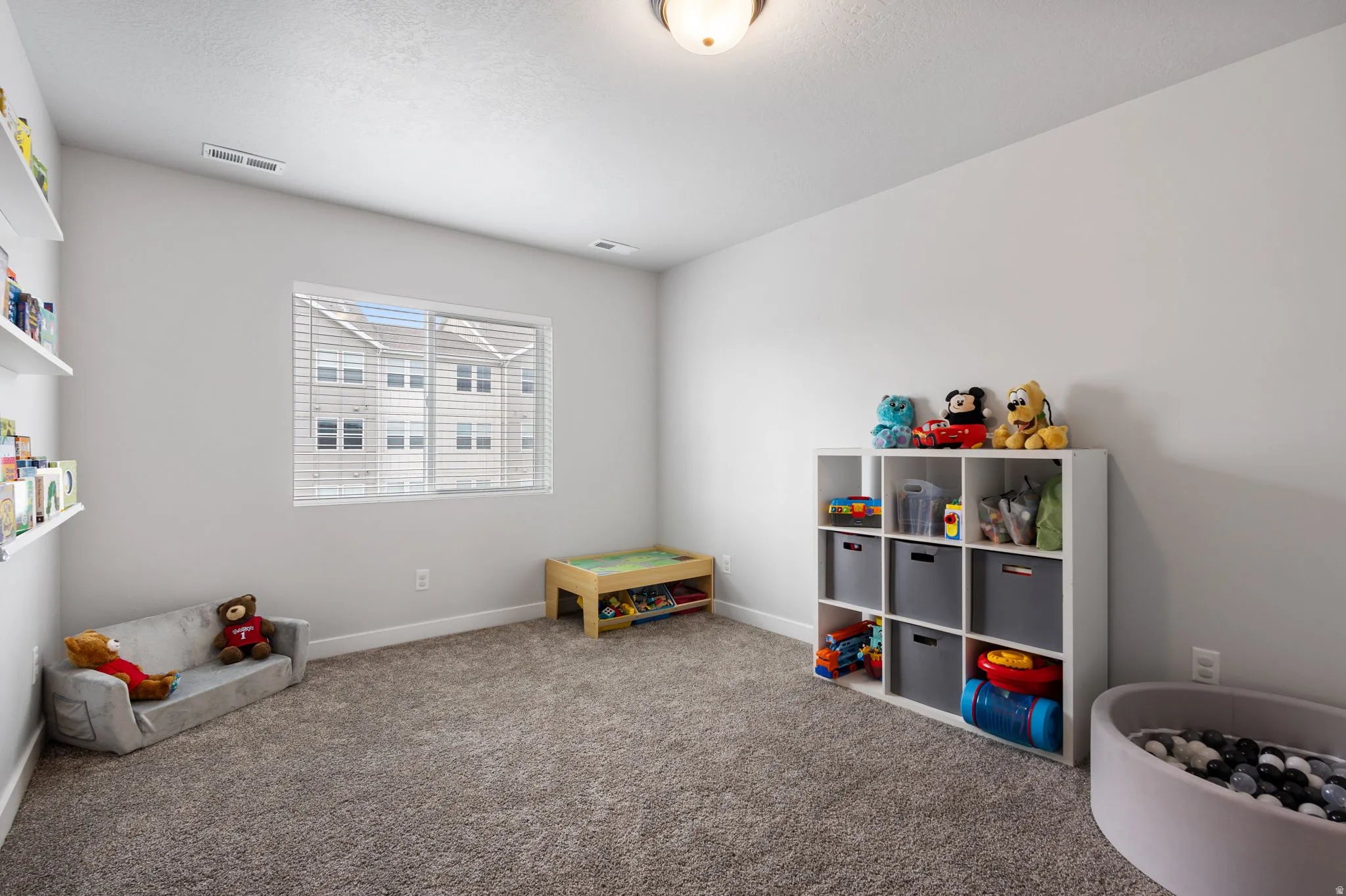 Carpeted room featuring a large window with blinds, overhead lighting, baseboard trim, and built-in wall shelving