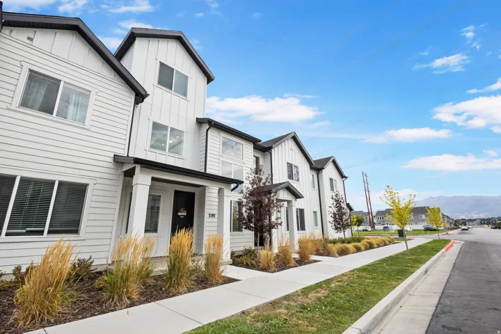 Modern townhome exterior featuring white siding with black trim, a covered entry porch, and mature landscaping