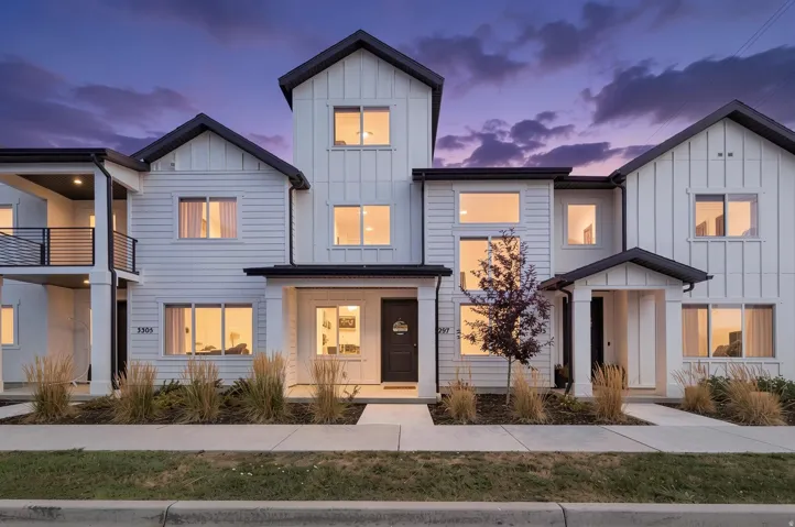Modern townhome exterior featuring white siding, prominent black-framed windows, dark roofing, a covered porch entry, and a second-story balcony with black railings