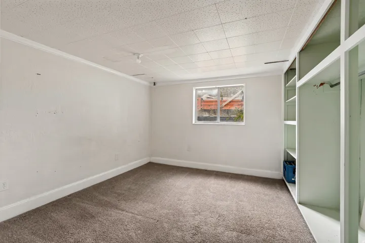 Carpeted room featuring a drop-panel ceiling, built-in shelving, a single window, white baseboards, and white crown molding