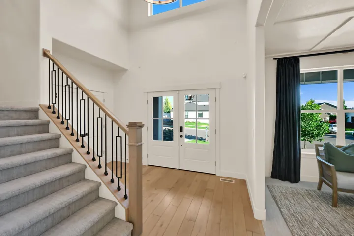 Two-story entryway with light wood-finish flooring, a grand staircase featuring light gray carpeted treads and black metal balusters, and a double-door entry with clear glass panels