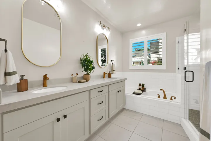 Primary Bathroom featuring a dual vanity with light cabinetry and solid surface countertop, an undermount tub with subway tile surround, a separate glass-enclosed shower, and a window with white louvered shutters