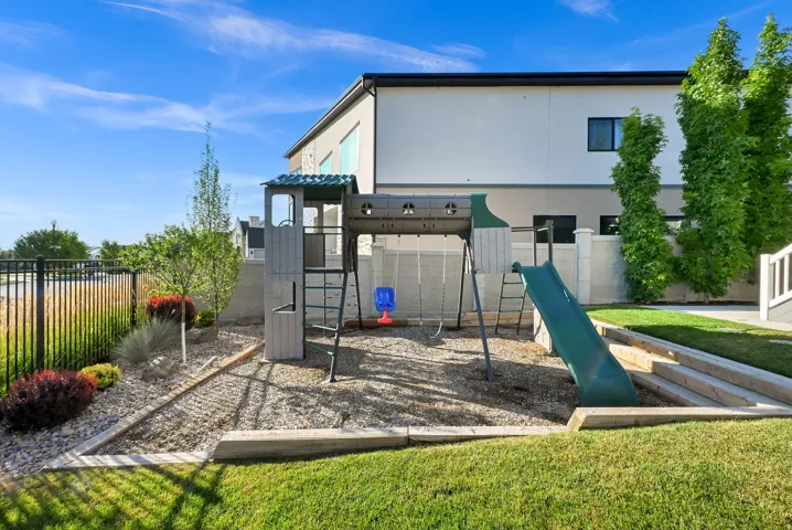 Backyard featuring a designated play area with pea gravel surfacing, a black metal fence, tiered landscaping with mature shrubs, and a manicured lawn