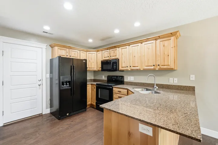 Kitchen featuring wood-finish flooring, a black appliance suite, light wood cabinetry, stone countertops, and a double basin sink with a high-arc faucet