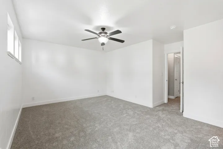 Spacious room featuring light gray carpeting, crisp white walls, a multi-pane window, ceiling fan with integrated lighting, and white baseboard trim