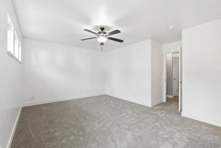 Spacious room featuring light gray carpeting, crisp white walls, a multi-pane window, ceiling fan with integrated lighting, and white baseboard trim