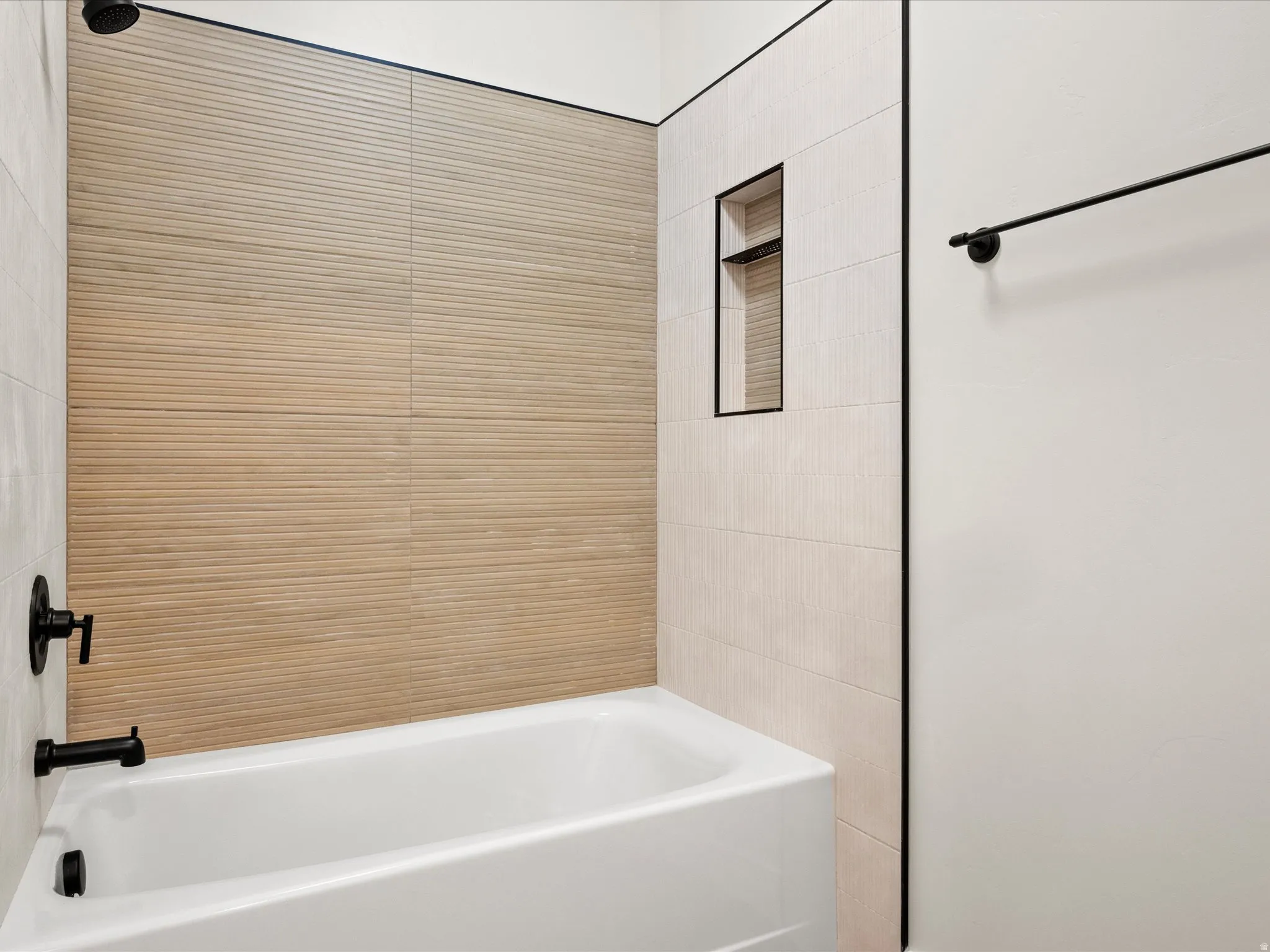 Bathtub with matte black fixtures, textured beige tile surround, light cream vertical tile accents, and a recessed shower niche