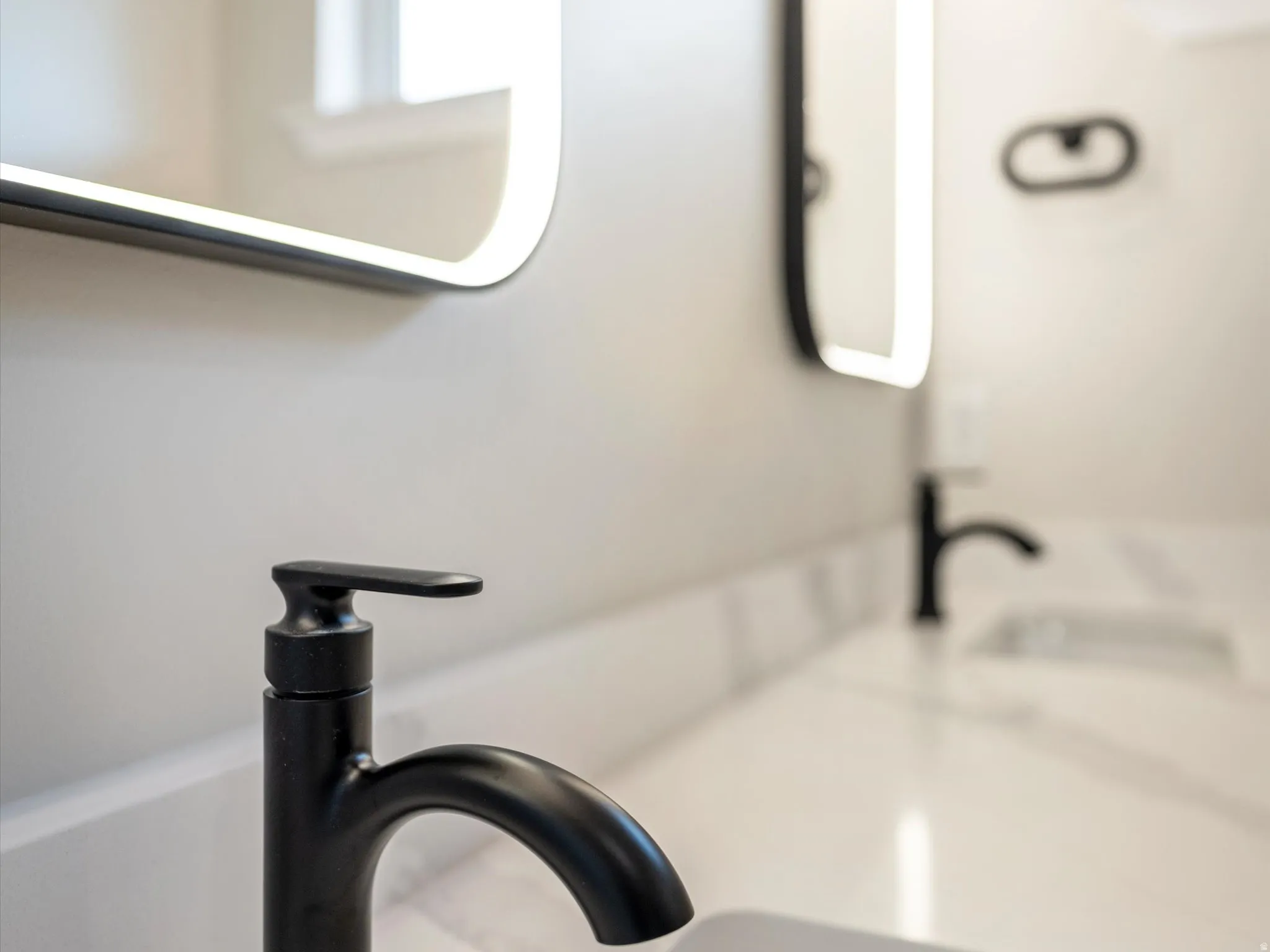 Contemporary bathroom vanity featuring dual matte black faucets, integrated sinks, a white countertop with subtle veining, and illuminated mirrors with rounded corners