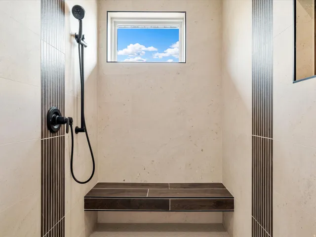 Walk-in shower with natural-toned stone tile walls, a built-in wood-finish bench, and a matte black shower fixture