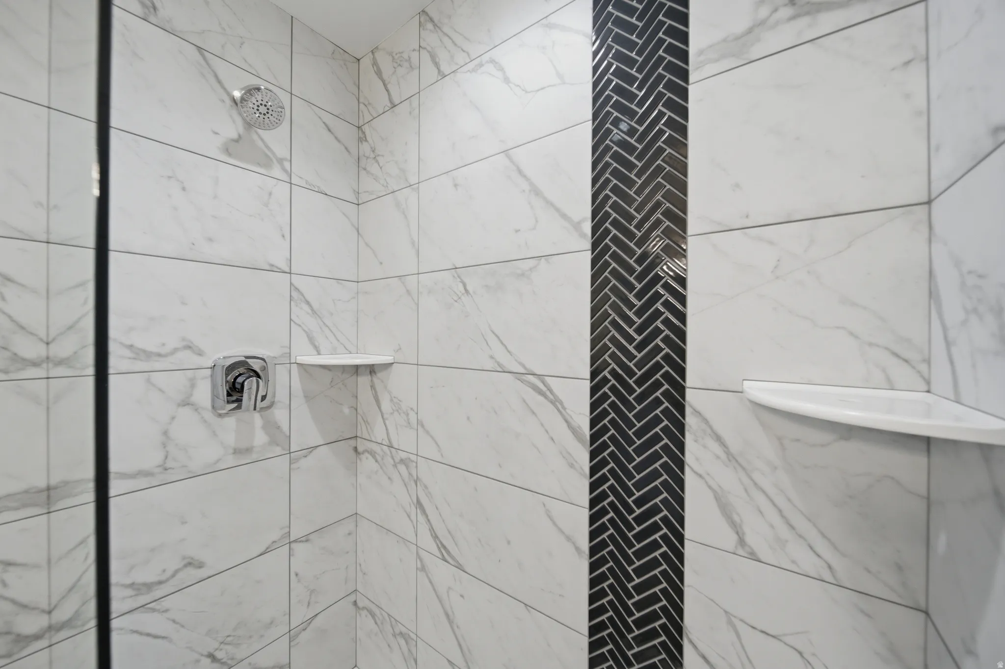 Shower enclosure featuring white marble-look tile, a black herringbone mosaic accent strip, chrome shower head, polished chrome mixer, and two built-in corner shelves