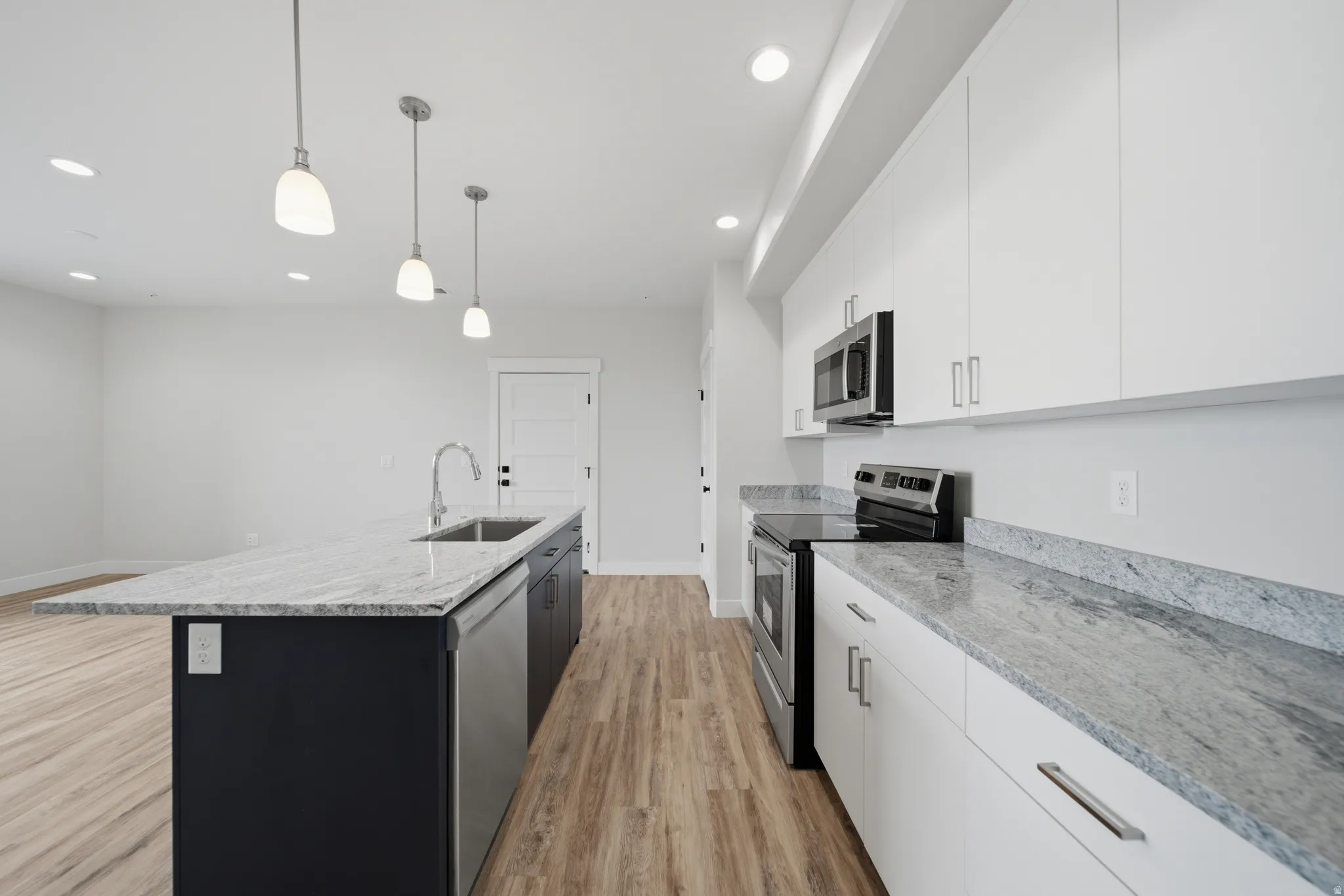 Modern kitchen featuring an expansive center island with an undermount sink, granite countertops, stainless steel appliances, white flat-panel cabinetry, and wood-finish flooring