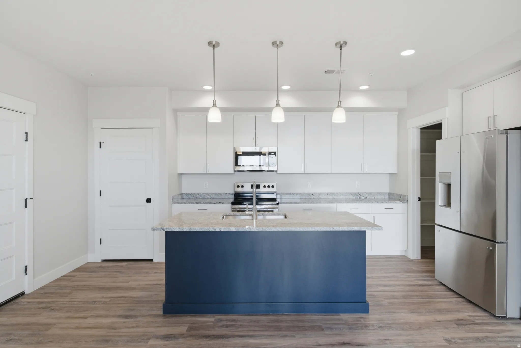 Kitchen featuring a blue-painted island with a light-toned countertop and sink, three pendant lights, white cabinetry, stainless steel appliances, and wood-finish flooring