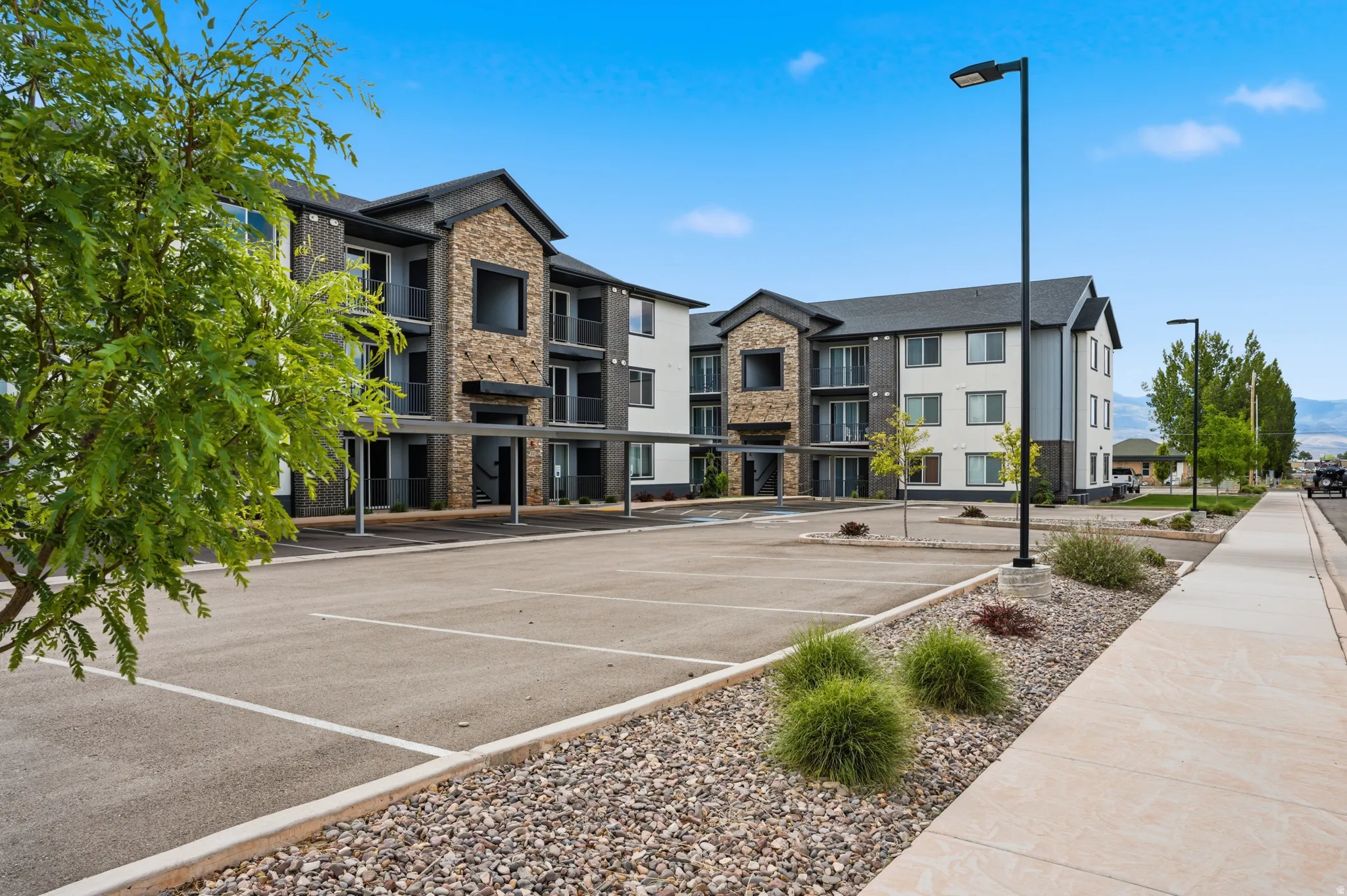 Multi-story building featuring a stone and dark siding facade, covered parking, individual unit balconies, a paved parking lot, and sidewalk access