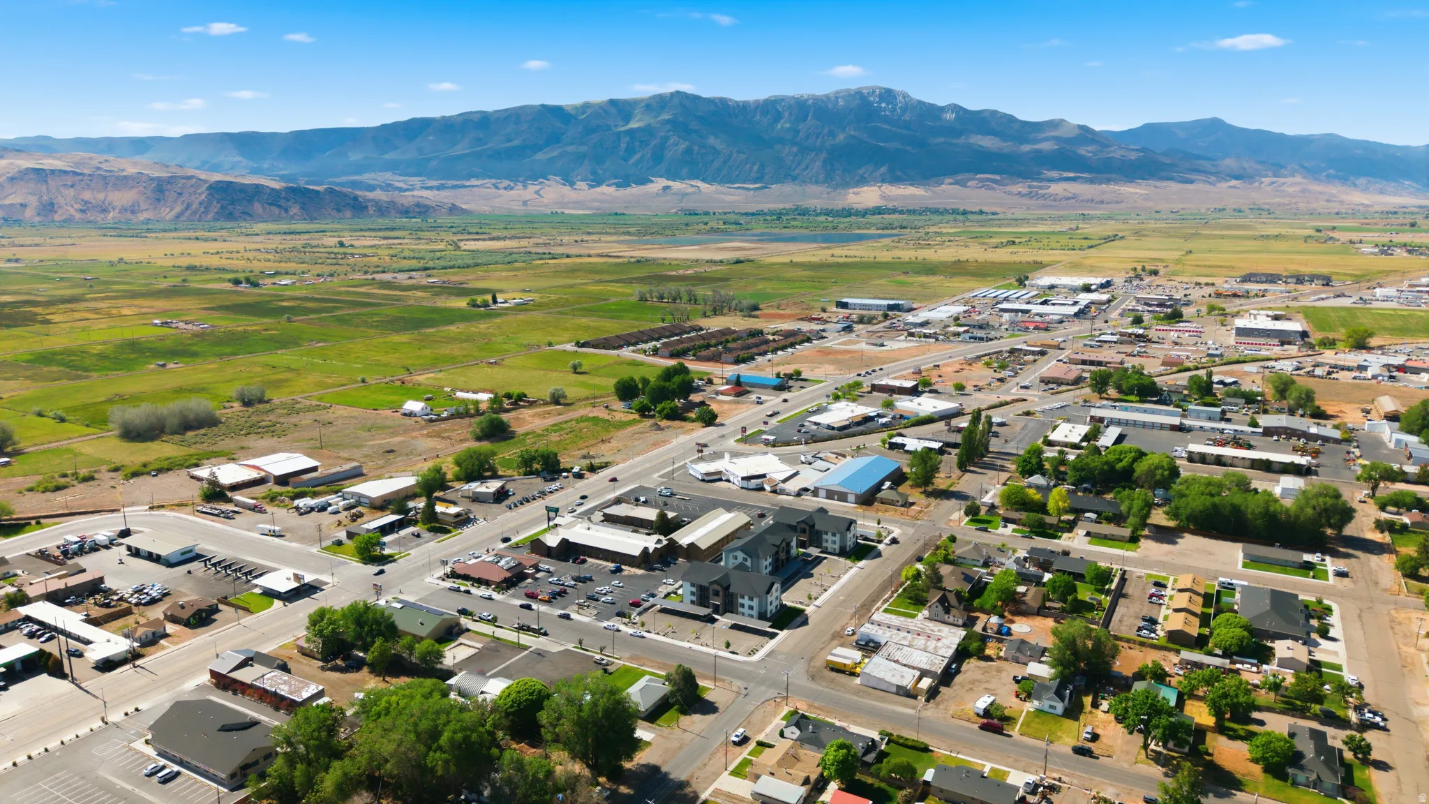 Expansive aerial perspective showcasing a town with varied commercial and residential structures, bordered by vast green fields and distant mountains