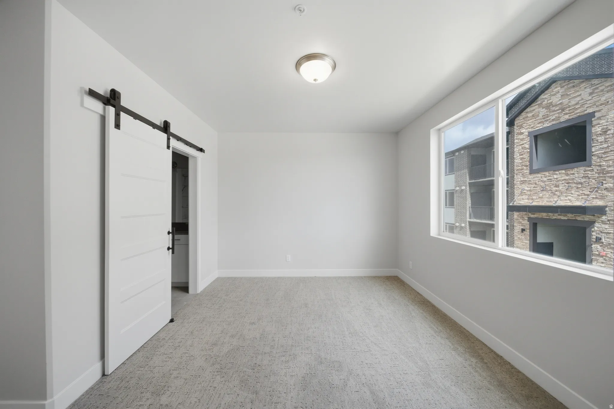 Spacious room featuring a white barn door, neutral-toned carpeting, white baseboards, and a large window providing natural light