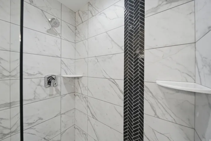 Shower enclosure featuring white marble-look tile, a black herringbone mosaic accent strip, chrome shower head, polished chrome mixer, and two built-in corner shelves