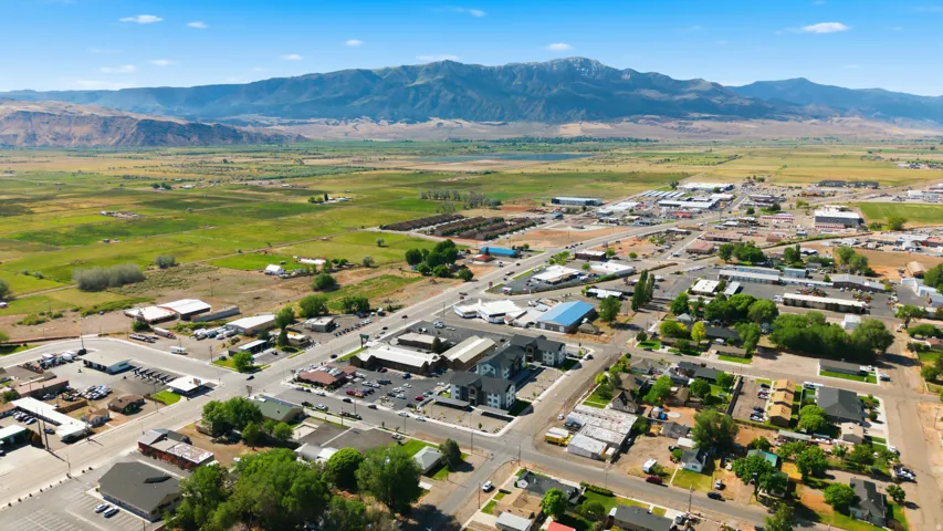 Expansive aerial perspective showcasing a town with varied commercial and residential structures, bordered by vast green fields and distant mountains
