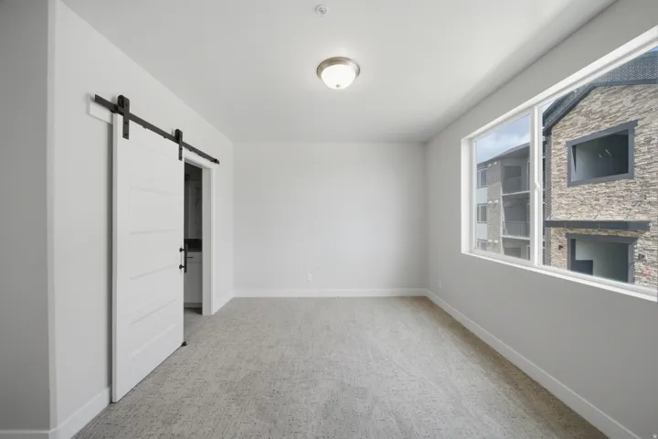 Spacious room featuring a white barn door, neutral-toned carpeting, white baseboards, and a large window providing natural light